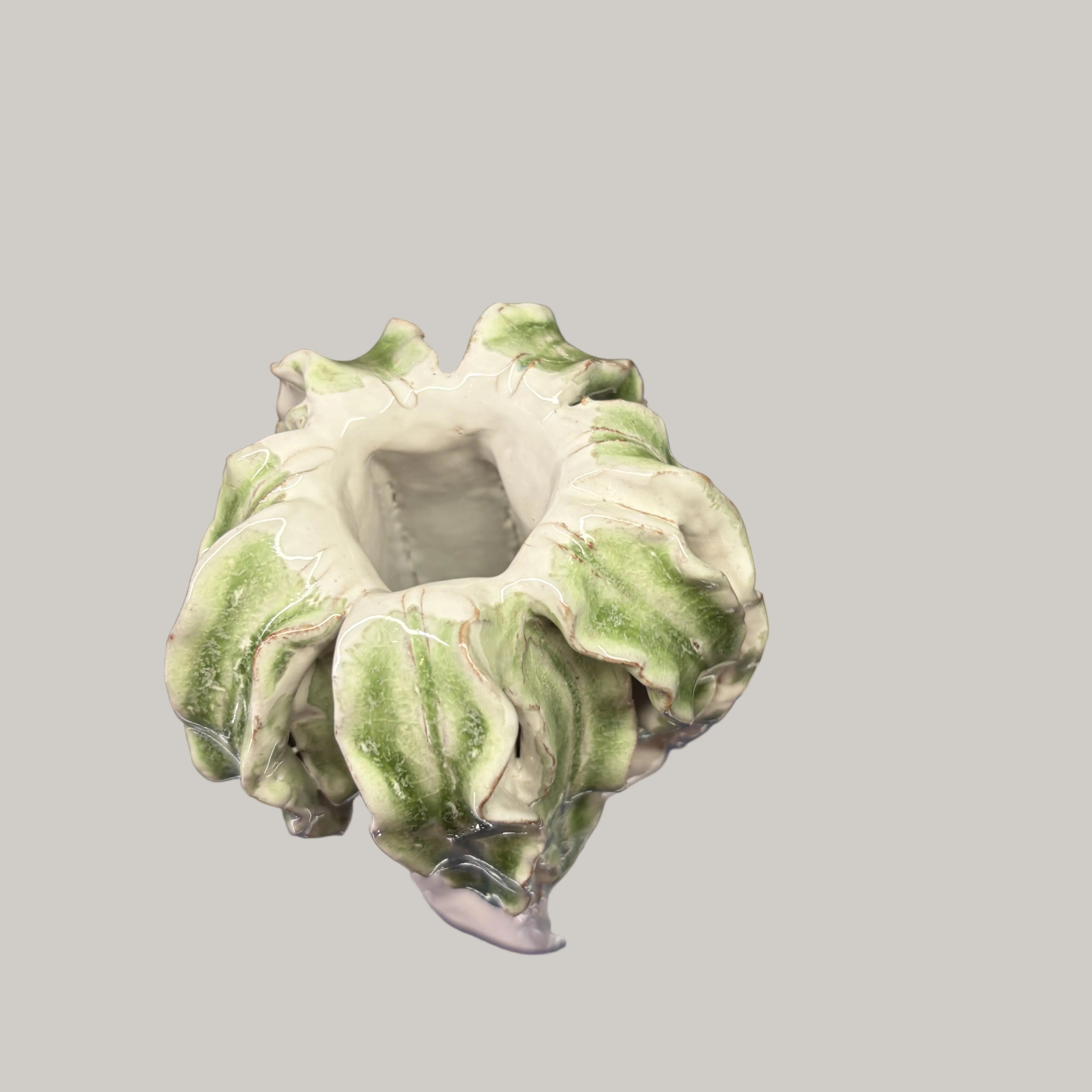 A small sculptural ceramic planter or vase with a ruffled, flower-like rim, shown wrapped in clear plastic on top of a blue book. The item has a white base with green marbled or variegated detailing around the outer edges.