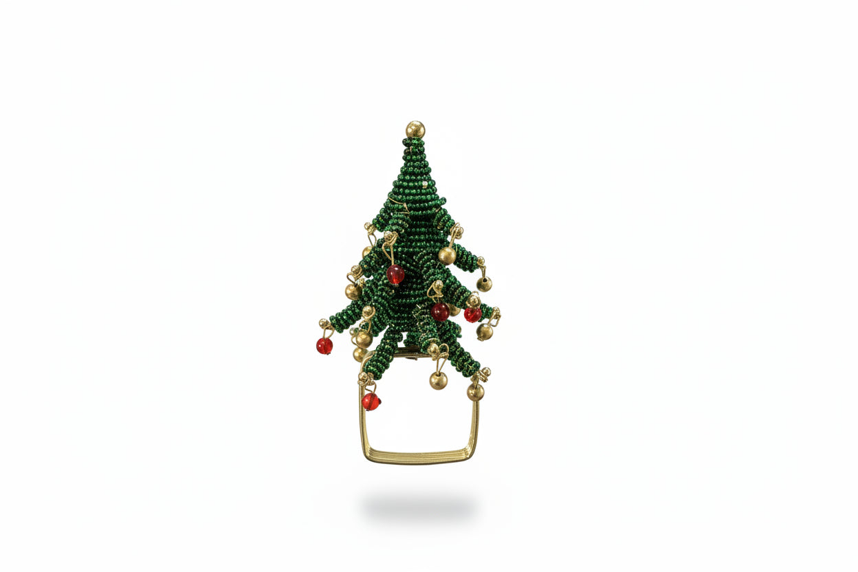 A hand holding a small green beaded Christmas tree ornament with gold and red accents, set against a background of a dark wooden dresser and gray floor.
