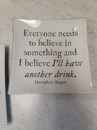 A square white ceramic plate with a glossy finish, featuring a black printed quote by Humphrey Bogart: 'Everyone needs to believe in something and I believe I'll have another drink.' The plate is photographed on a light gray marble surface.