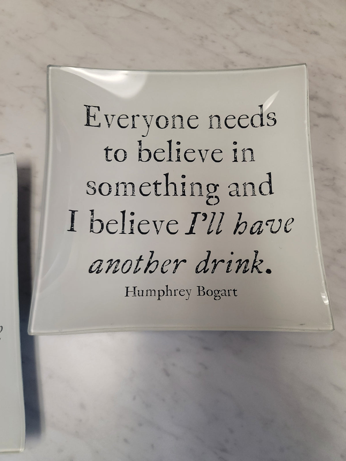 A square white ceramic plate with a glossy finish, featuring a black printed quote by Humphrey Bogart: 'Everyone needs to believe in something and I believe I'll have another drink.' The plate is photographed on a light gray marble surface.