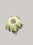 A small sculptural ceramic planter or vase with a ruffled, flower-like rim, shown wrapped in clear plastic on top of a blue book. The item has a white base with green marbled or variegated detailing around the outer edges.