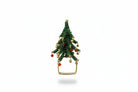 A hand holding a small green beaded Christmas tree ornament with gold and red accents, set against a background of a dark wooden dresser and gray floor.